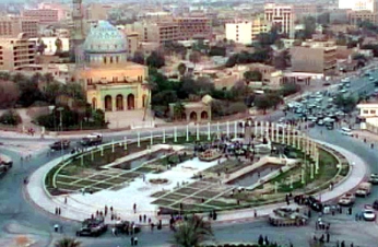 A long shot of Firdos Square during the statue toppling process. A small knot of onlookers can be seen surrounding the statue at the far end of the area; most of the square is empty. Three US tanks can be seen stationed around the square.