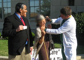 Health reform organizer Randall Terry pretends to stab an elderly lady in the neck as part of an anti-reform protest. A fellow protester wearing a Barack Obama mask looks on.